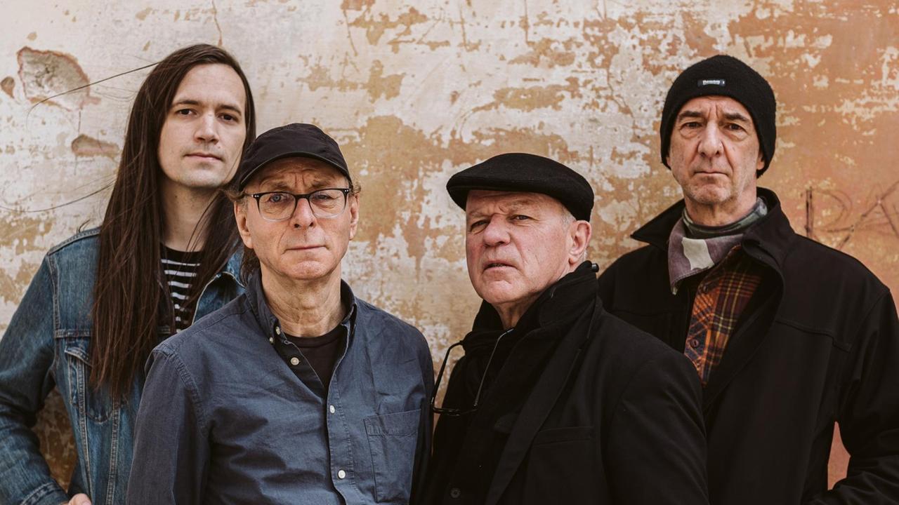 Vier Männer stehen vor einer okerfarbenen Wand. Zusammen sind sie die Band Wire. Vier Männer stehen vor einer okerfarbenen Wand. Zusammen sind sie die Band Wire.