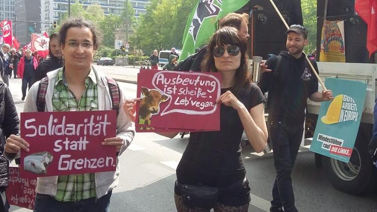 Eine junge Frau im schwarzen T-Shirt, mit Sonnenbrille nimmt an einem sonnigen Tag auf einem belebten Platz an einer Demonstration teil. Auf dem Schild. das sie hochhält ist eine Kuh mit einer Sprechblase zu sehen, in der geschrieben steht: "Ausbeutung ist scheiße. Leb' vegan." Eine junge Frau im schwarzen T-Shirt, mit Sonnenbrille nimmt an einem sonnigen Tag auf einem belebten Platz an einer Demonstration teil. Auf dem Schild. das sie hochhält ist eine Kuh mit einer Sprechblase zu sehen, in der geschrieben steht: "Ausbeutung ist scheiße. Leb' vegan."