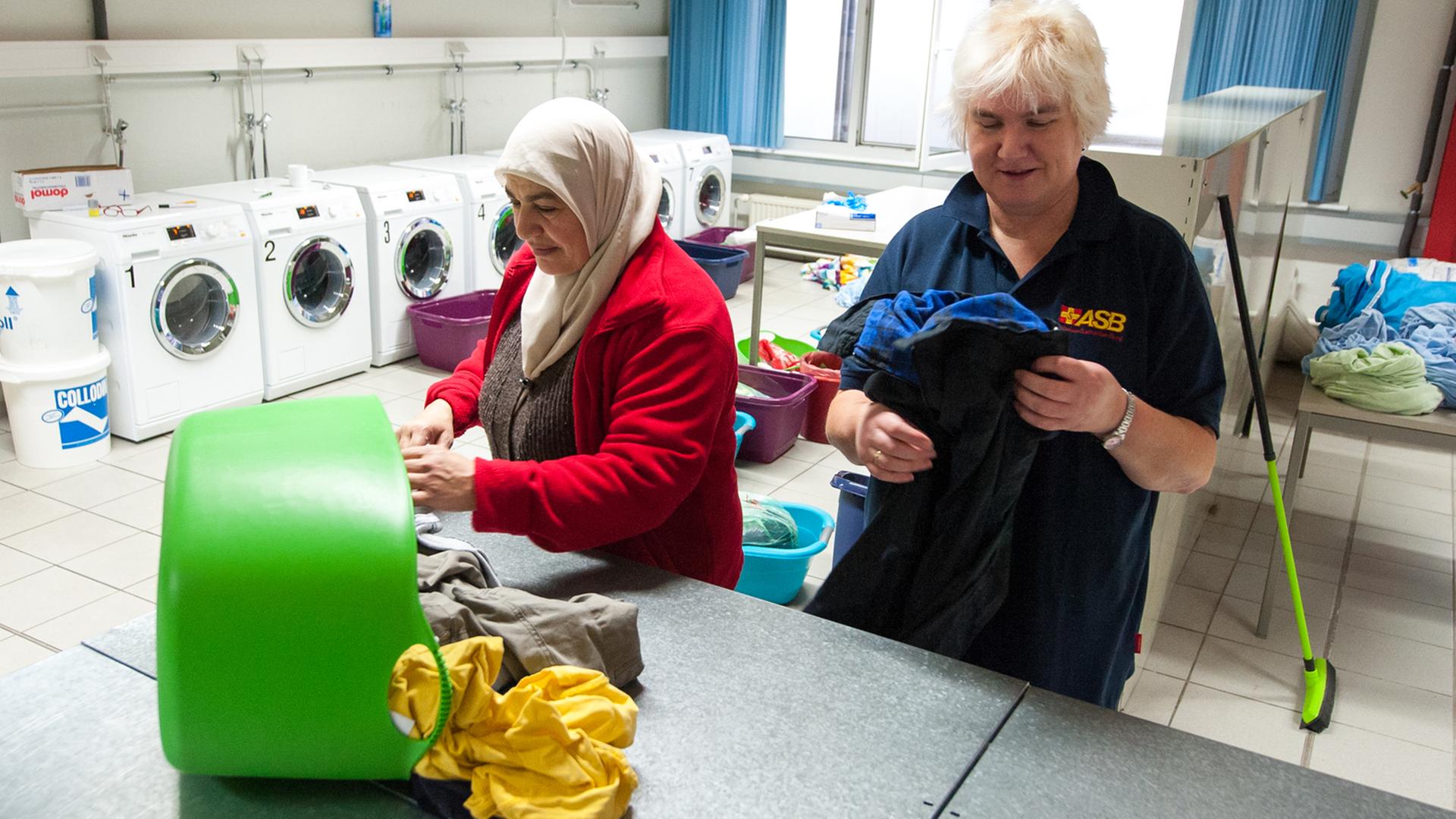 Eine Dame legt zusammen mit einer Flüchtlingsfrau Kleidung in einer Wäscherei zusammen.