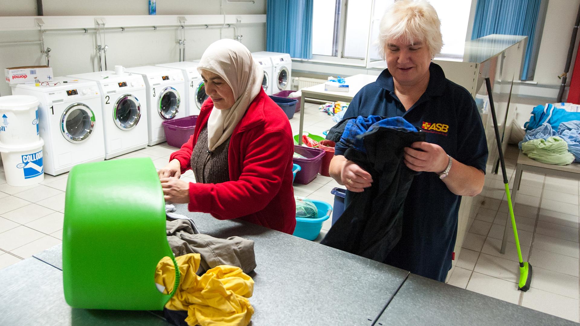 Eine Dame legt zusammen mit einer Flüchtlingsfrau Kleidung in einer Wäscherei zusammen. Eine Dame legt zusammen mit einer Flüchtlingsfrau Kleidung in einer Wäscherei zusammen.