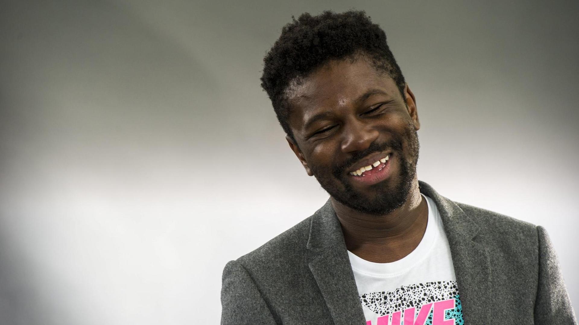Michael Donkor kam in London in einem ghanaischen Haushalt zur Welt. Auf dem Foto trägt er ein graues Sakko und ein T-Shirt mit pinkem Nike-Schriftzug und Logo. Michael Donkor kam in London in einem ghanaischen Haushalt zur Welt. Auf dem Foto trägt er ein graues Sakko und ein T-Shirt mit pinkem Nike-Schriftzug und Logo.