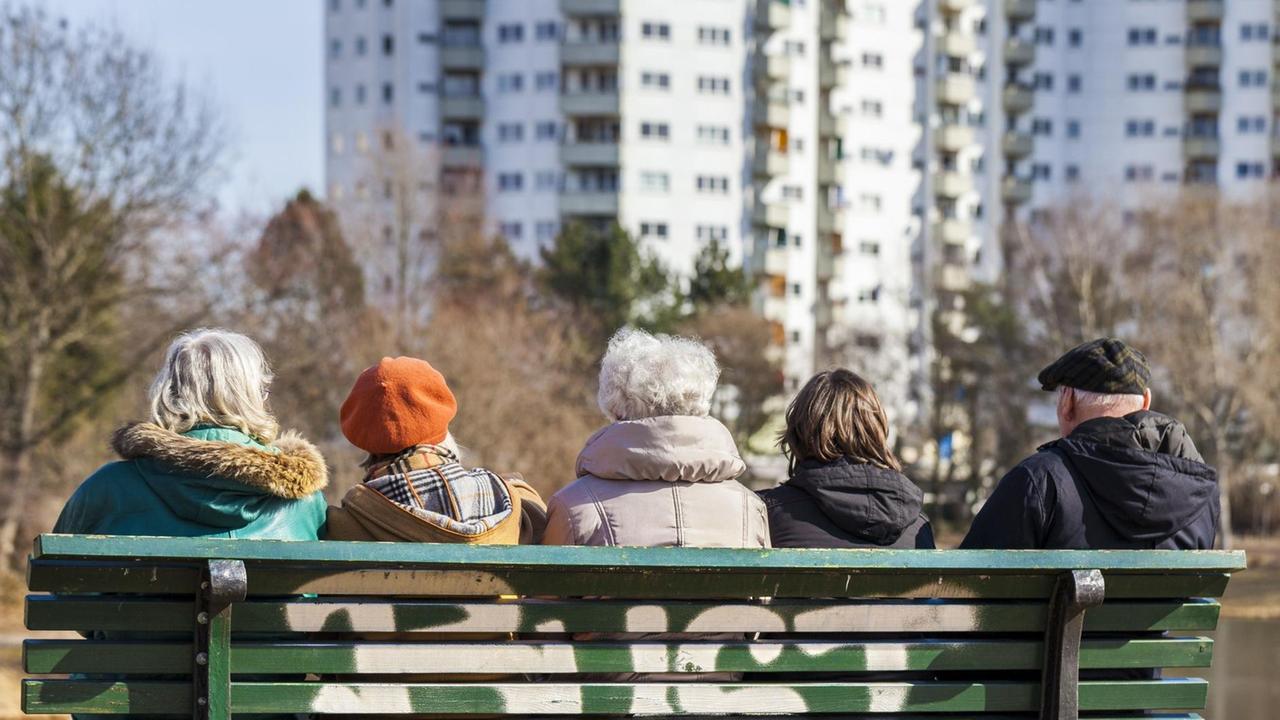 Mehrere Senioren sitzen auf einer Bank, im Hintergrund ist ein großer Wohnungsblock zu sehen. Mehrere Senioren sitzen auf einer Bank, im Hintergrund ist ein großer Wohnungsblock zu sehen.