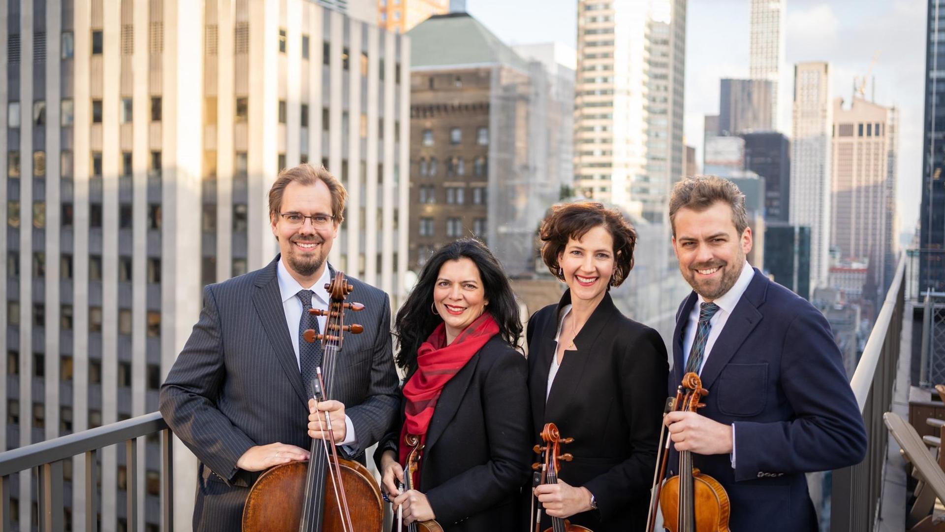 Die vier Ensemblemitglieder stehen mit ihren Instrumenten auf einem Balkon, hinter dem die Skyline von New York mit blauem Himmel zu sehen ist. Die vier Ensemblemitglieder stehen mit ihren Instrumenten auf einem Balkon, hinter dem die Skyline von New York mit blauem Himmel zu sehen ist.