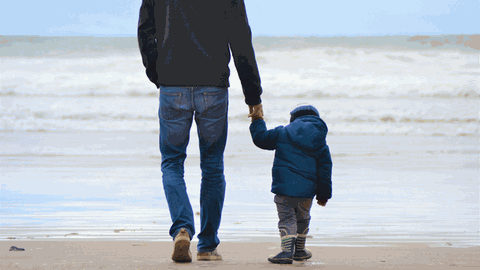 En Mann geht mit einem kleinen Kind an der Hand an einem Strand auf das Meer zu.