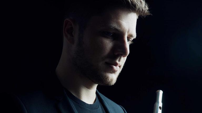 Der Musiker Matvey Demin ist mit seiner Flöte im Profil und im Halbdunkel zu sehen. Der Musiker Matvey Demin ist mit seiner Flöte im Profil und im Halbdunkel zu sehen.