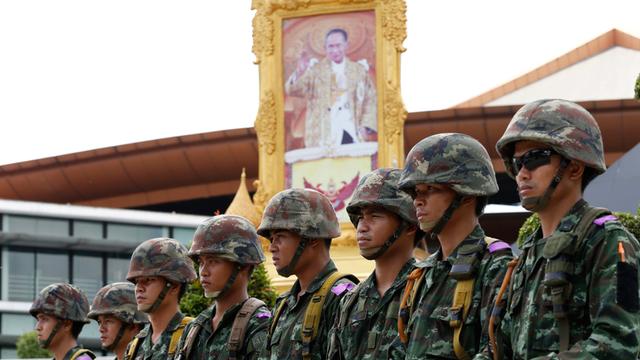 Eine Reihe von acht uniformierten Soldaten steht mit abgesetzten Schutzschilden vor einem Porträt von König Bhumibol Adulyadej