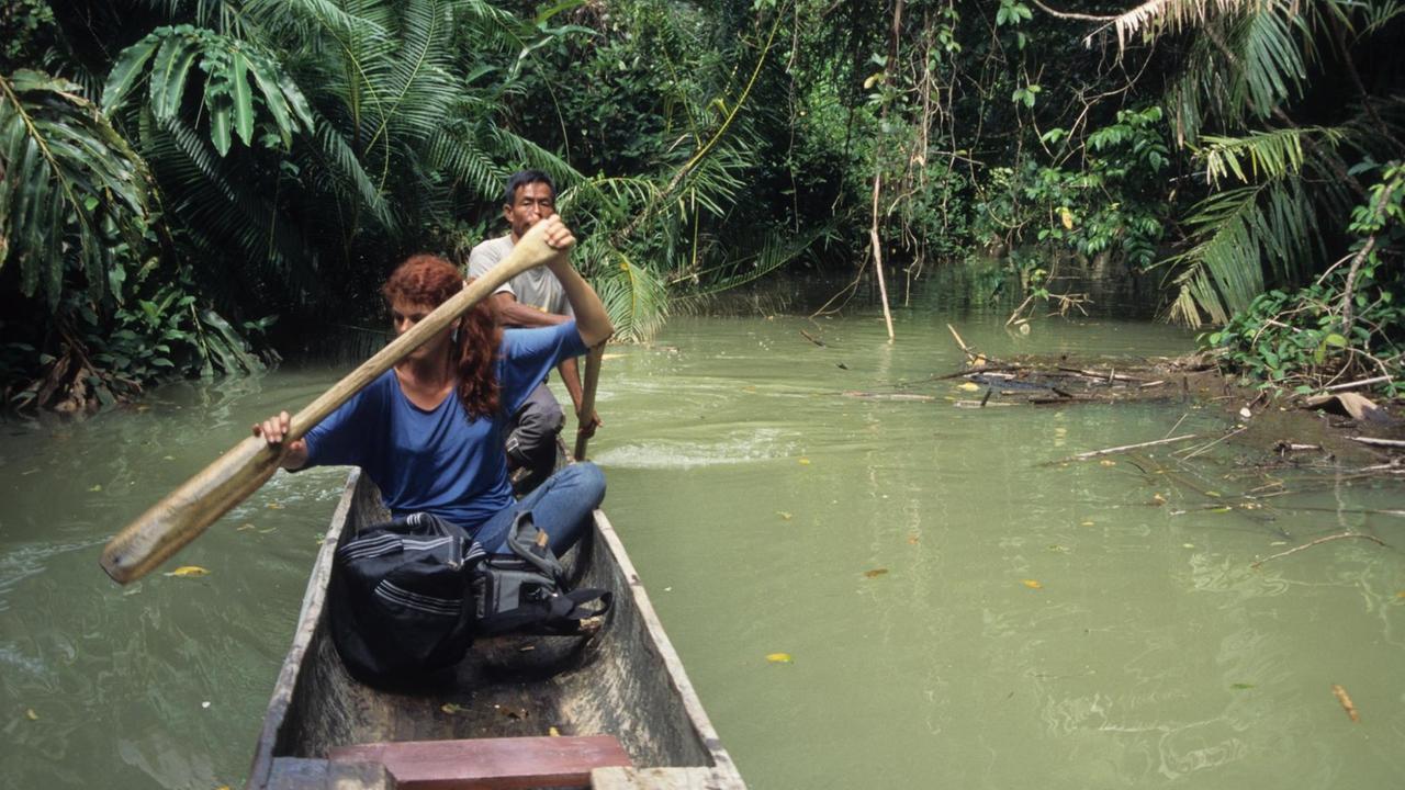 Eine Frau mit langen, roten Haaren rudert in einem Holzboot auf einem Urwaldfluss. Eine Frau mit langen, roten Haaren rudert in einem Holzboot auf einem Urwaldfluss.