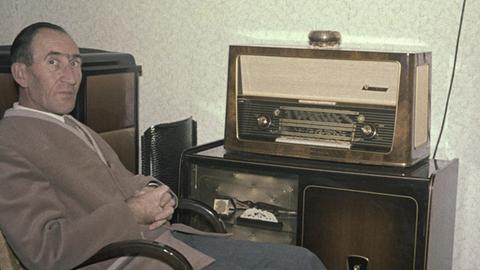 Ein Mann sitzt auf einem historischen Foto in einem Schaukelstuhl und hört Radio.