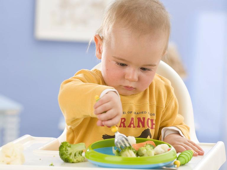 Ein Baby sitzt in einem Hochstuhl, hat einen Teller mit Gemüsestücken vor sich und sticht mit seiner Gabel in ein Stück Brokkoli.