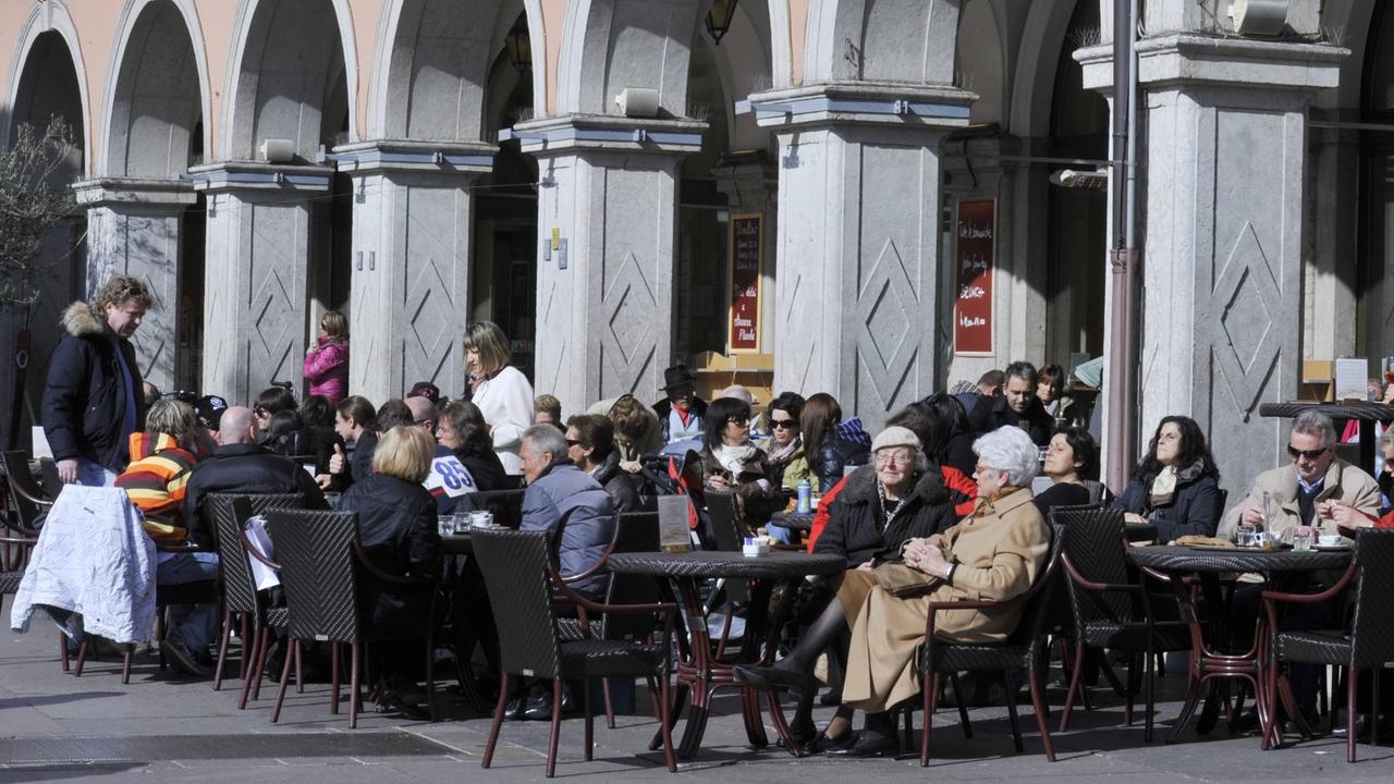 Menschen sitzen in einem Straßencafé in Bozen. Im Hintergrund ein Säulengang.