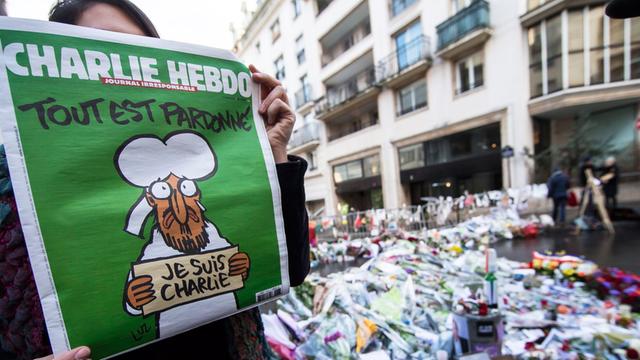Eine Frau zeigt die Zeitschrift "Charlie Hebdo". Im Hintergrund liegen Blumen für die Opfer der Anschläge auf die Redaktion.