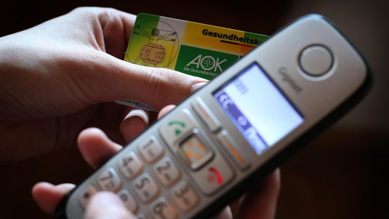 Ein Mann hält eine Gesundheitskarte der Allgemeinen Ortskrankenkasse (AOK) in der Hand, während er am Telefon eine Nummer wählt. Ein Mann hält eine Gesundheitskarte der Allgemeinen Ortskrankenkasse (AOK) in der Hand, während er am Telefon eine Nummer wählt.