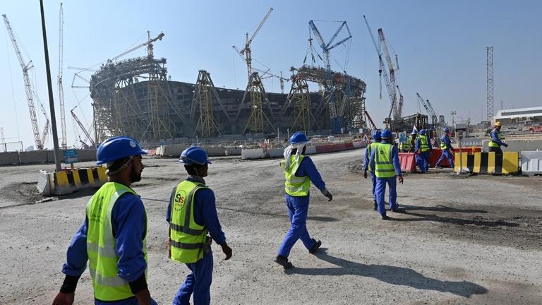 Männer in Arbeitskleidung gehen vor der Baustelle eines Stadions entlang. Männer in Arbeitskleidung gehen vor der Baustelle eines Stadions entlang.