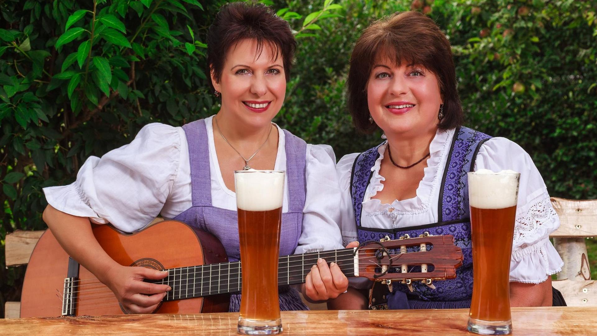 Zwei Frauen im Dirndl sitzen im Garten an einem Tisch, musizieren und singen zusammen. Zwei Frauen im Dirndl sitzen im Garten an einem Tisch, musizieren und singen zusammen.