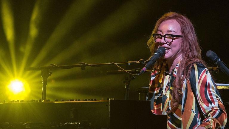 Tori Amos sitzt im farbenfrohen Kleid am Flügel, links im Hintergrund leuchtet ein Scheinwerfer strahlenförmig in Gelb. Tori Amos sitzt im farbenfrohen Kleid am Flügel, links im Hintergrund leuchtet ein Scheinwerfer strahlenförmig in Gelb.