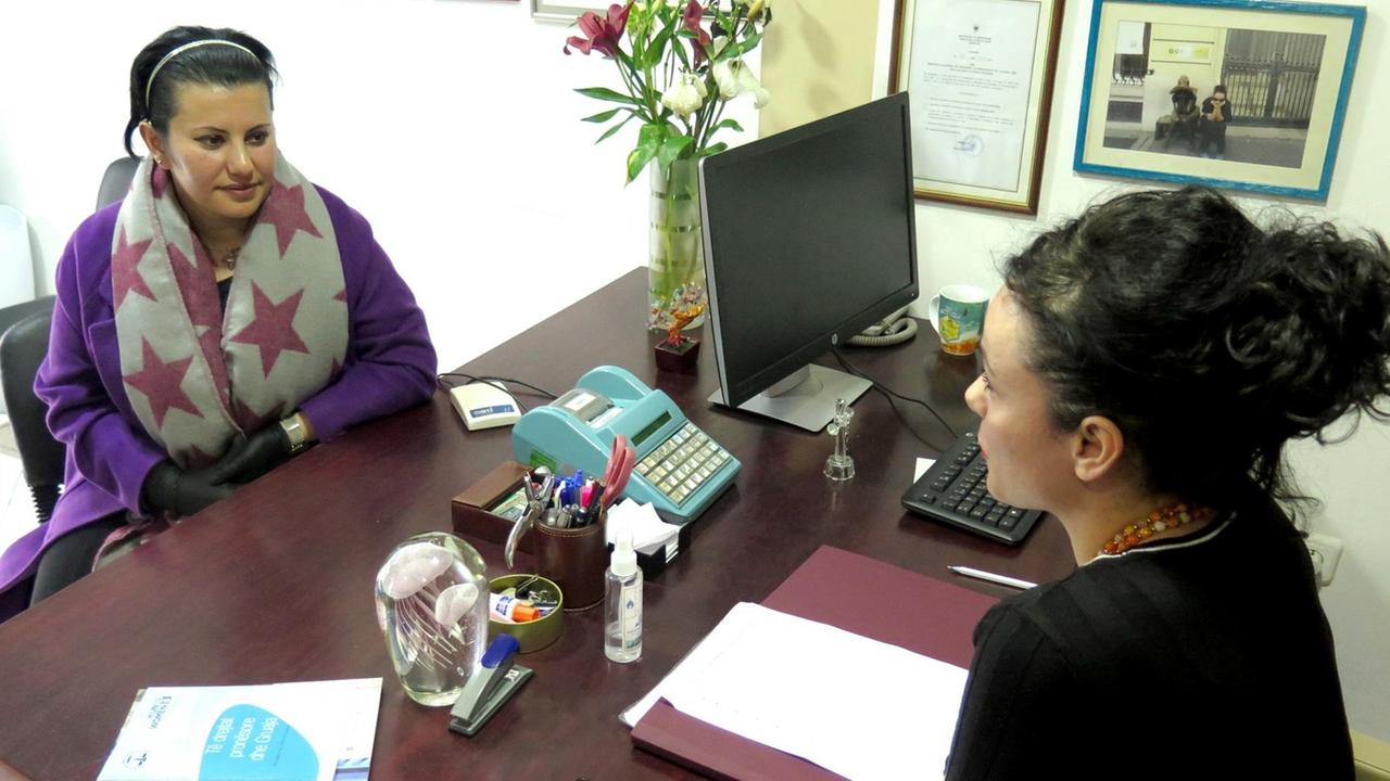 Ilda Cadra (l.), in lila Mantel und großem Schal mit roten Sternen,  und die Juristin Elona Saliaj im schwarzen Pulli sitzen sich an einem Schreibtisch in einem Büro gegenüber.