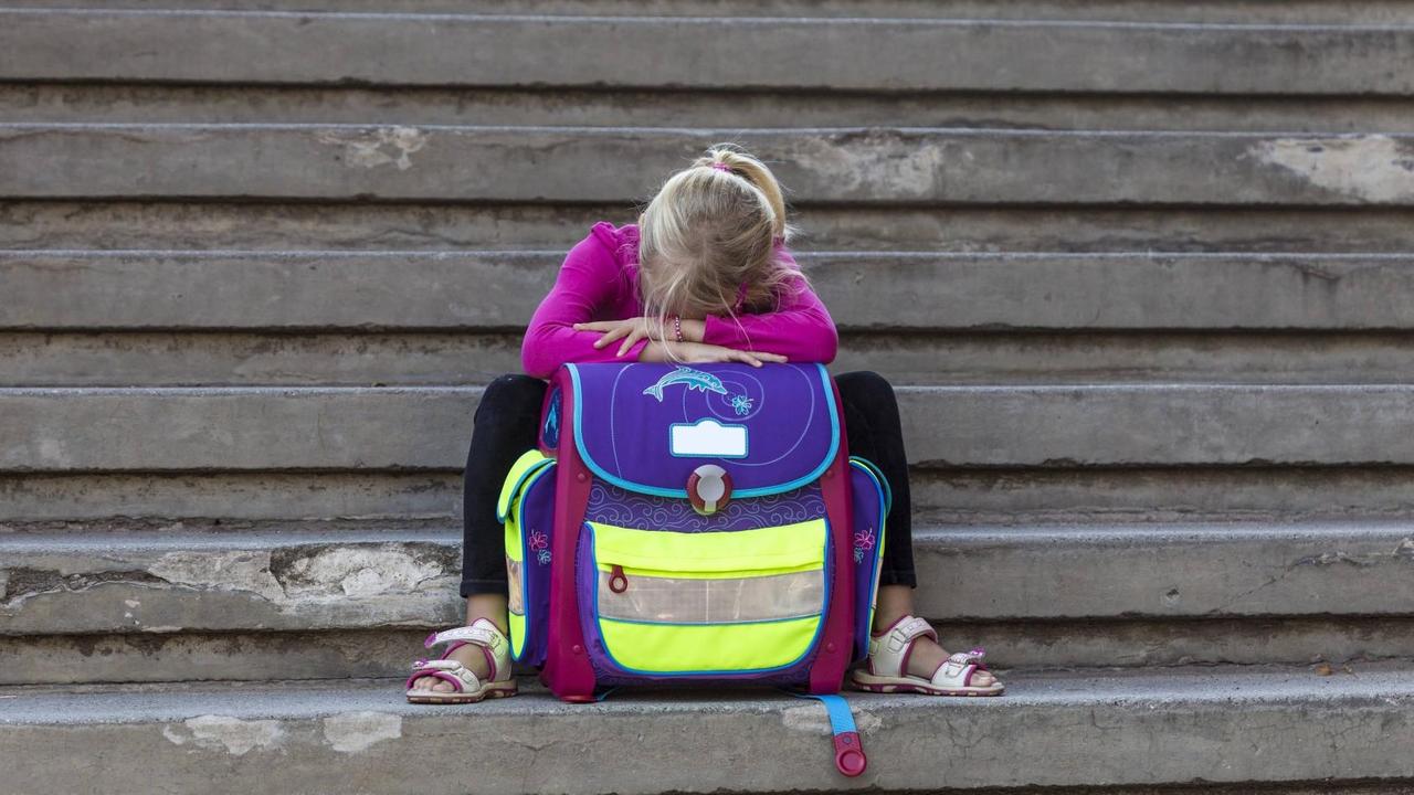Ein kleines Mädchen sitzt auf Stufen, den Kopf in ihren Schulranzen vergraben. Ein kleines Mädchen sitzt auf Stufen, den Kopf in ihren Schulranzen vergraben.