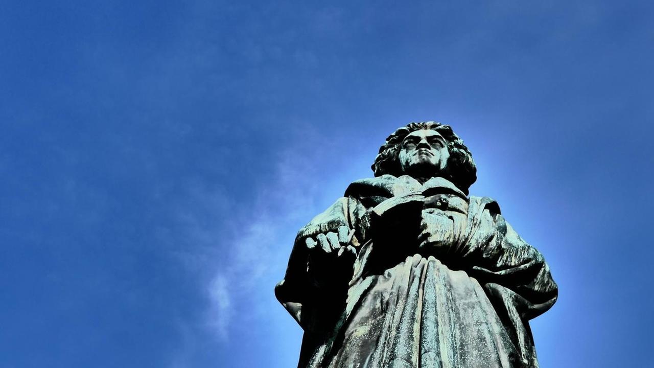 Blick hinauf am Beethovendenkmal in Bonn am Rhein, wo die grau-grüne Bronzefigur in den blauen Himmel ragt. Blick hinauf am Beethovendenkmal in Bonn am Rhein, wo die grau-grüne Bronzefigur in den blauen Himmel ragt.