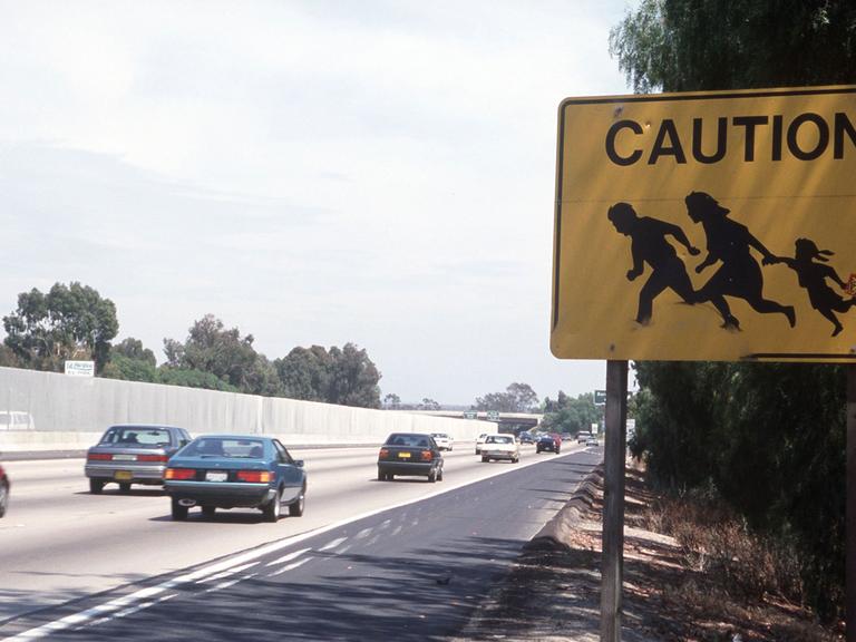 Ein Verkehrsschild an einer stark befahrenen Straße entlang der Grenze zwischen San Diego in den USA und Tijuana in Mexiko mahnt zur Vorsicht vor flüchtenden Mexikanern, die auf ihrem Weg in die Staaten plötzlich die Straße überqueren könnten und ein Unfallrisiko darstellen.
