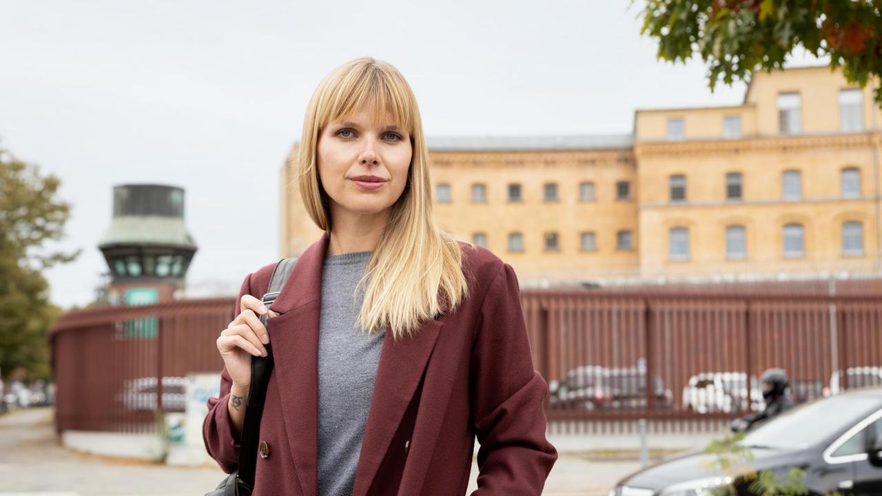 Ein junge Frau mit langen, glatten blonden Haaren und weinrotem Blazer steht vor einem Gefängnisgebäude.