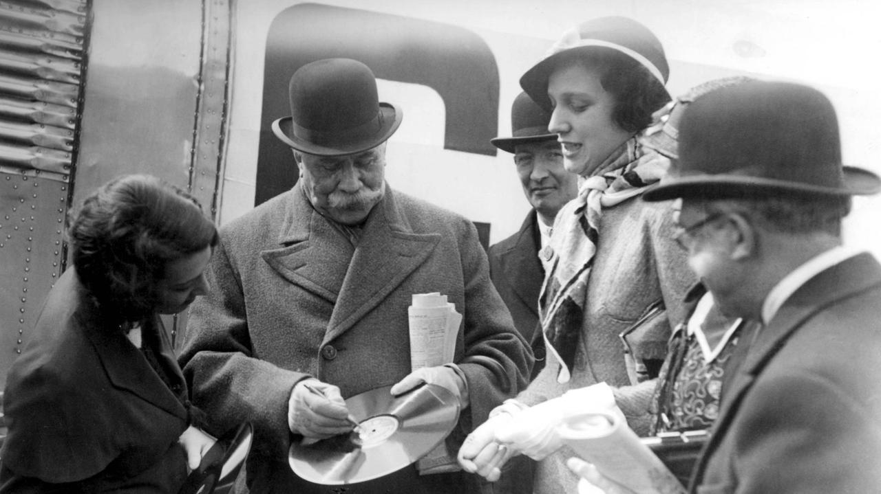 Der Komponist mit Melone und Mantel vor einem Flugzeug ist von vier weiteren Personen umkreist und schauen zu, wie er eine Platte signiert.
