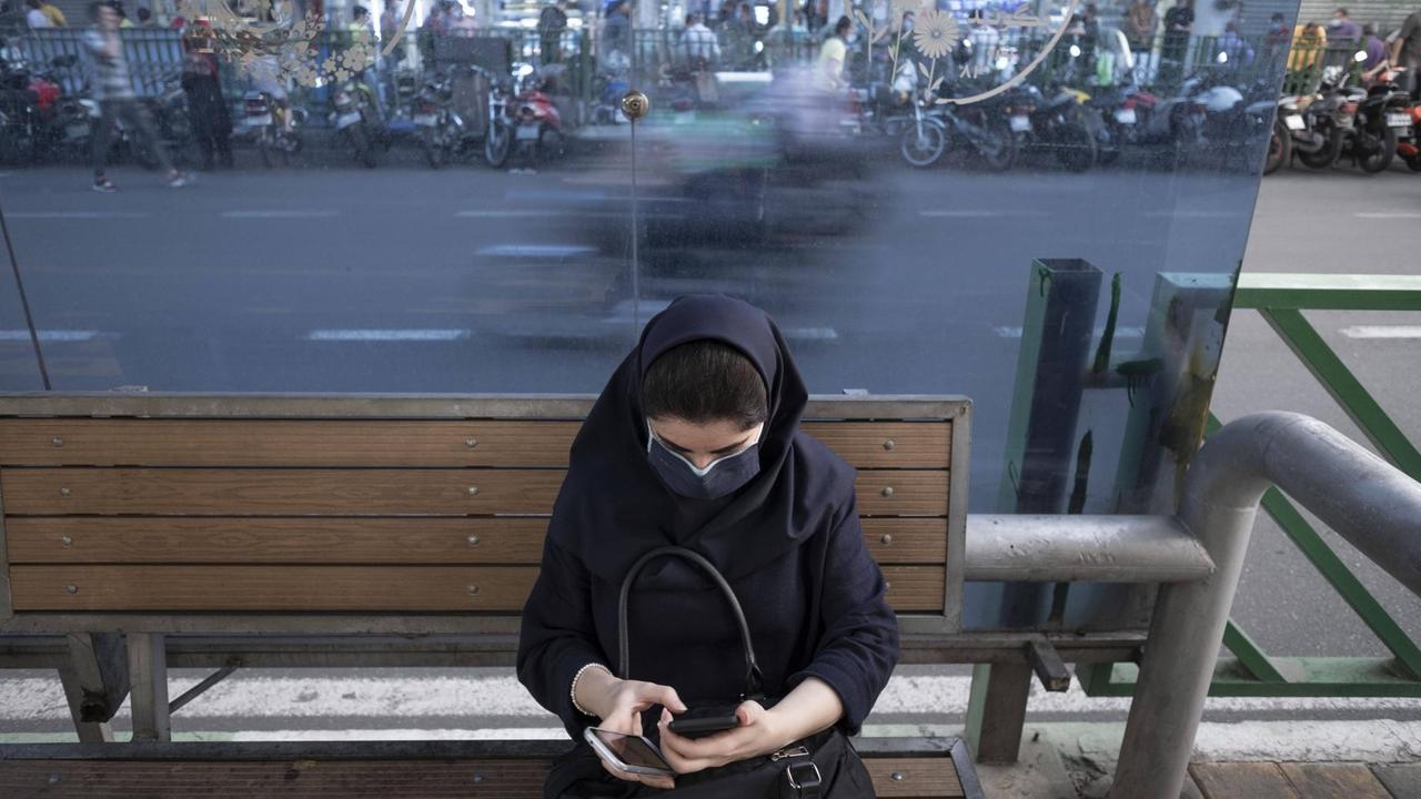 Ein Frau mit dunklem Kopftuch, dunkler Jacke und Mund-Nasen-Schutz sitzt auf einer Bank an einer Straße und schaut auf das Display eines Smartphones. Ein Frau mit dunklem Kopftuch, dunkler Jacke und Mund-Nasen-Schutz sitzt auf einer Bank an einer Straße und schaut auf das Display eines Smartphones.