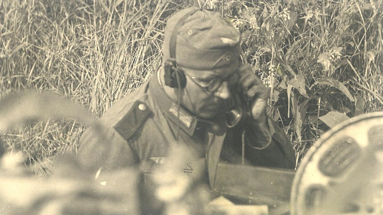 Schwarzweißfoto eines Mannes in Uniform, der im Gras liegt und Kopfhörer trägt. Schwarzweißfoto eines Mannes in Uniform, der im Gras liegt und Kopfhörer trägt.