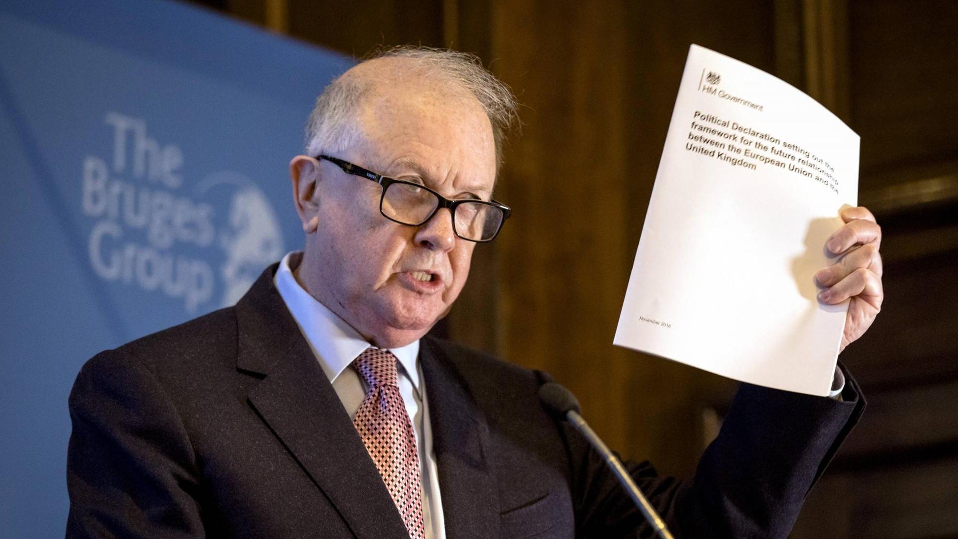 Barry Legg, der Vorsitzende der Bruges Group, steht an einem Pult unt hält ein Papier hoch. Barry Legg, der Vorsitzende der Bruges Group, steht an einem Pult unt hält ein Papier hoch.