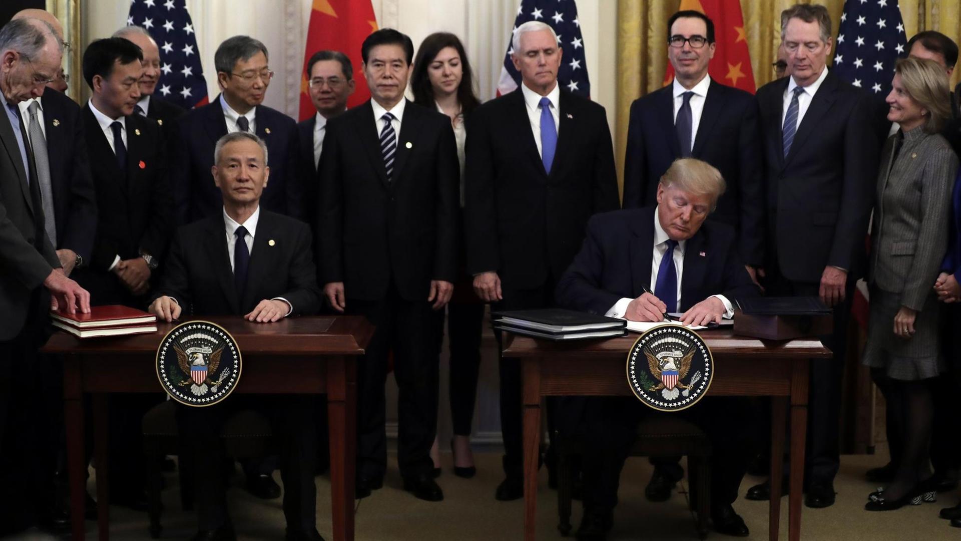 Donald Trump und Liu He unterzeichen an getrennten Tischen nebeneinander sitzend einen Vetrag. Im Hintergrund sind ihre Delegationen sowie die chinesische und die US-Flagge zu sehen.
