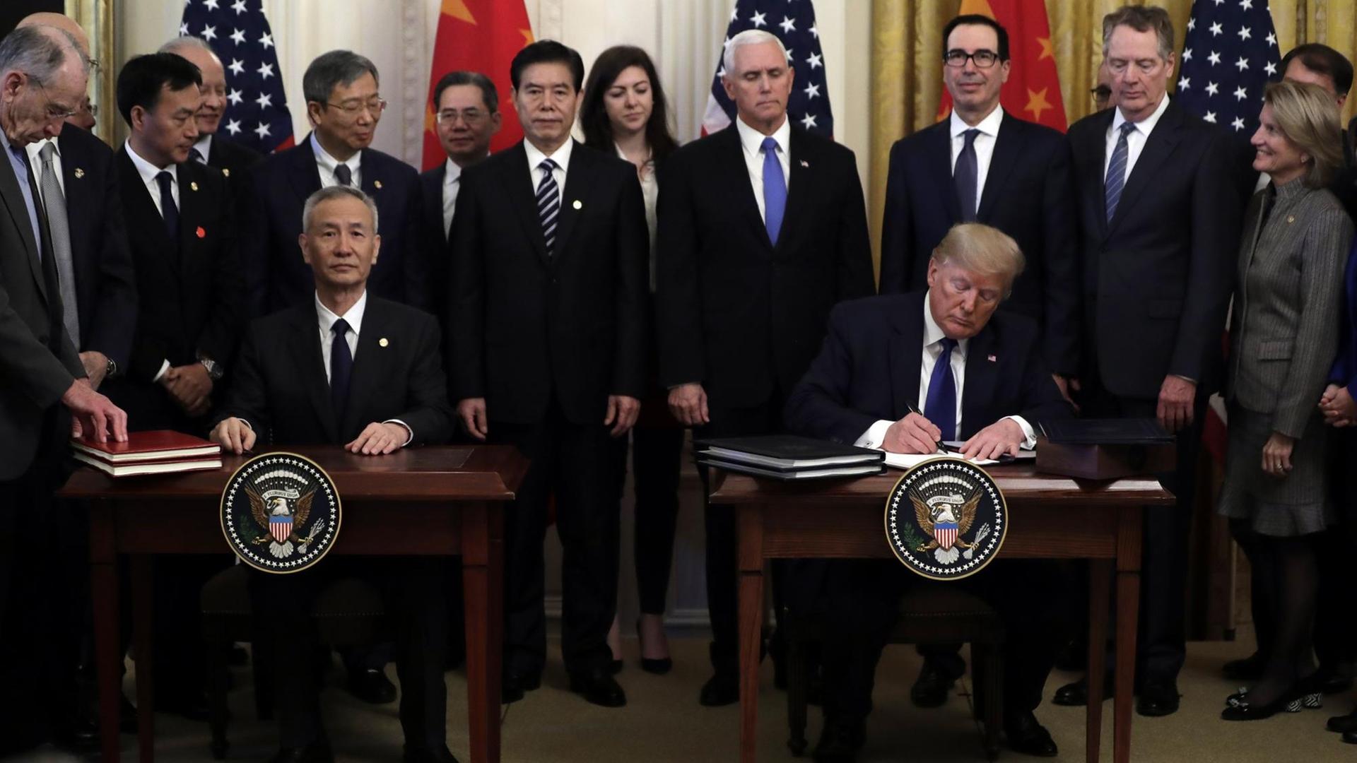 Donald Trump und Liu He unterzeichen an getrennten Tischen nebeneinander sitzend einen Vetrag. Im Hintergrund sind ihre Delegationen sowie die chinesische und die US-Flagge zu sehen. Donald Trump und Liu He unterzeichen an getrennten Tischen nebeneinander sitzend einen Vetrag. Im Hintergrund sind ihre Delegationen sowie die chinesische und die US-Flagge zu sehen.