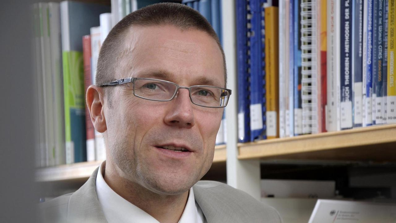 Ein Mann mit kurzen Haaren, Brille und grauem Anzug sitzt vor einem Bücherregal. Ein Mann mit kurzen Haaren, Brille und grauem Anzug sitzt vor einem Bücherregal.
