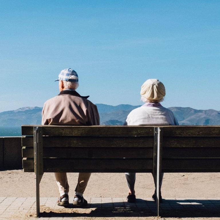 Zwei ältere Menschen sitzen auf einer Bank bei einer Aussichtsplattform und schauen auf die Berge.
