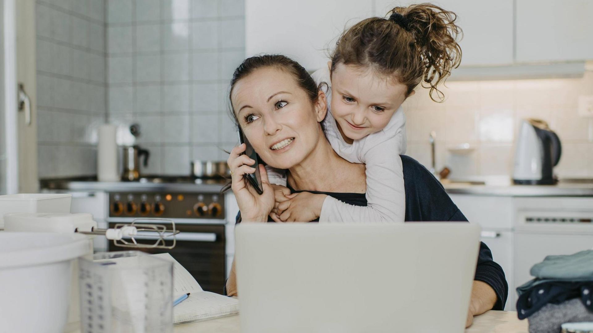 Mutter bei Homeoffice in der Küche, mit Kind am Hals und Haushalt um sich herum. Sie telefoniert und hat einen Laptop vor sich am Küchentisch.(Symbolbild) Mutter bei Homeoffice in der Küche, mit Kind am Hals und Haushalt um sich herum. Sie telefoniert und hat einen Laptop vor sich am Küchentisch.(Symbolbild)