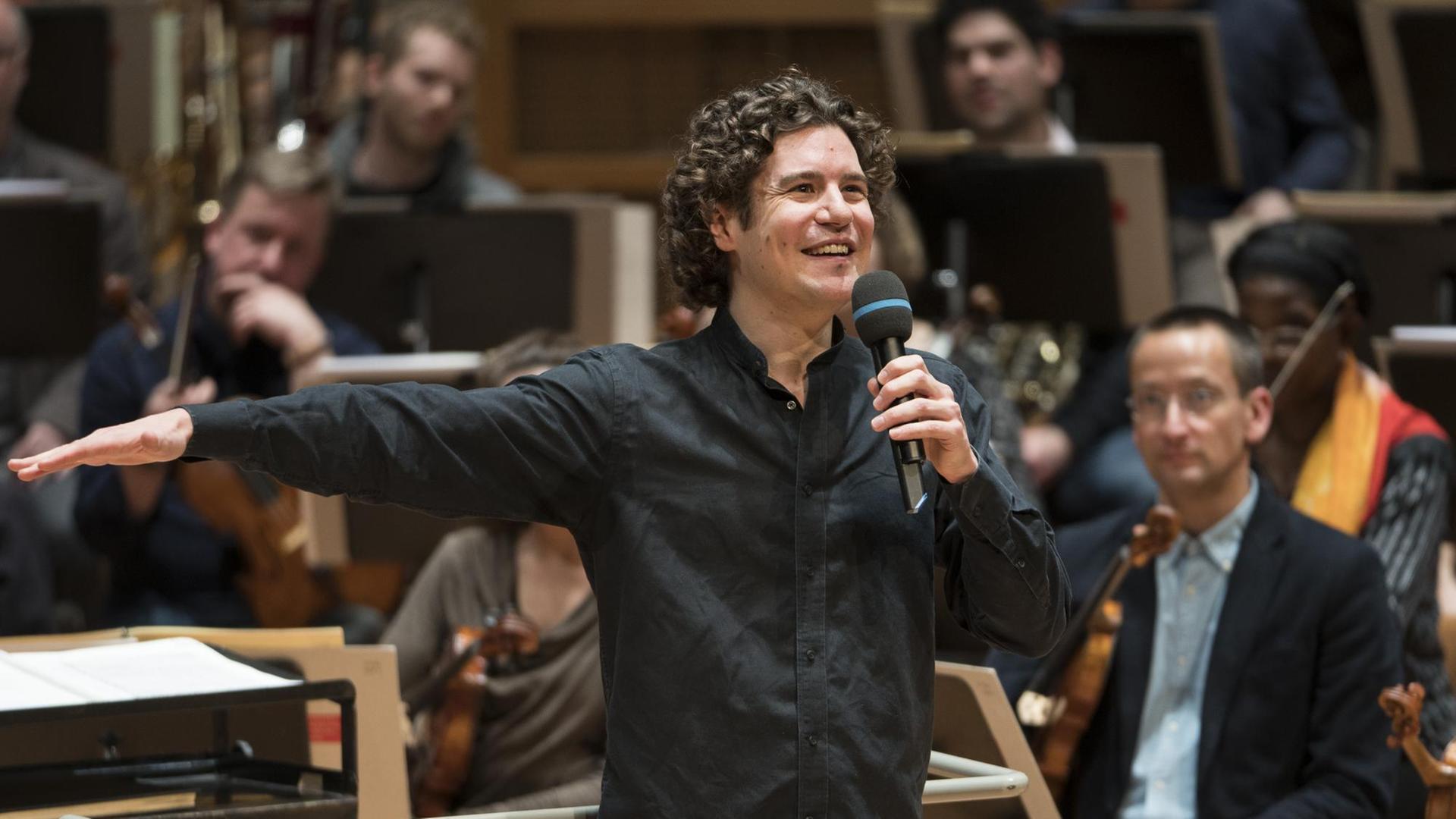 Der Dirigent Robin Ticciati steht vor dem Orchester, dem Deutschen Symphonie-Orchester Berlin, mit einem Mikrofon in der Hand und blickt lachend ins Publikum. Der Dirigent Robin Ticciati steht vor dem Orchester, dem Deutschen Symphonie-Orchester Berlin, mit einem Mikrofon in der Hand und blickt lachend ins Publikum.