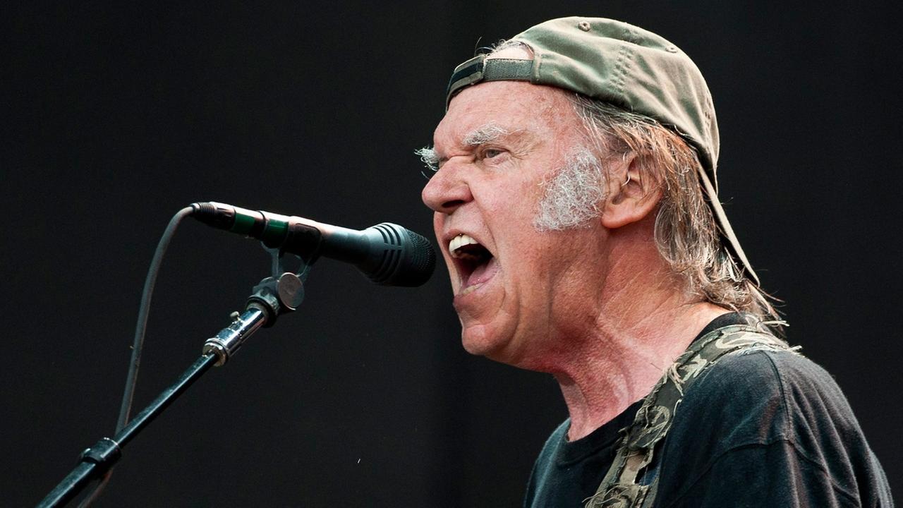 Ein Mann mit umgedrehter Baseballmütze und Gitarrengurt singt in ein Mikrofon. Ein Mann mit umgedrehter Baseballmütze und Gitarrengurt singt in ein Mikrofon.