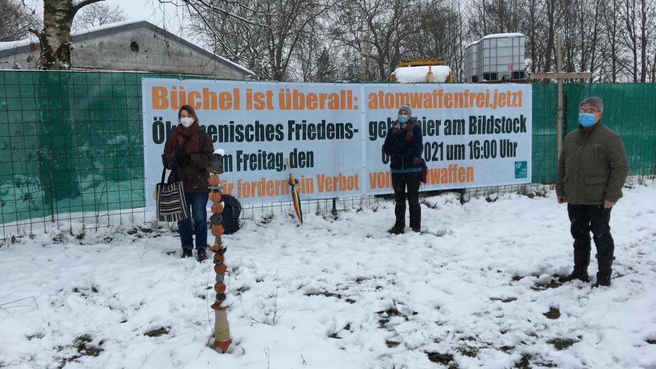 Menschen demonstrieren in Büchel auf einer verschneiten Wiese. Auf einem Transparent im Hintergrund steht: "Büchel ist überall - atomwaffenfrei jetzt. Ökumenisches Friedensgebiet hier am Bildstock. Am Freitag, den um 16 Uhr. Wir fordern ein Verbot von Atomwaffen" Menschen demonstrieren in Büchel auf einer verschneiten Wiese. Auf einem Transparent im Hintergrund steht: "Büchel ist überall - atomwaffenfrei jetzt. Ökumenisches Friedensgebiet hier am Bildstock. Am Freitag, den um 16 Uhr. Wir fordern ein Verbot von Atomwaffen"