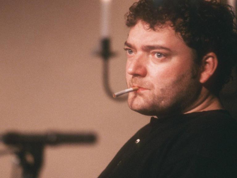 Wiglaf Droste sitzt auf einer Bühne mit Zigarette im Mund. Im Hintergrund steht ein Mikrofon.