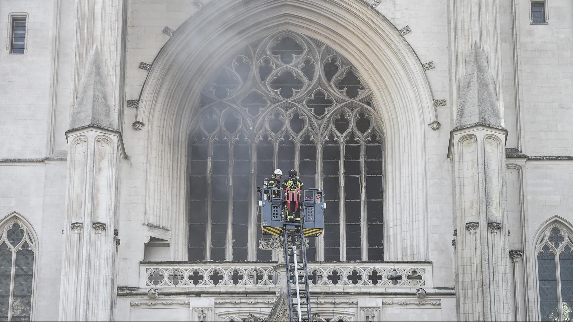 Feuerwehr-Leute stehen auf einer Leiter vor einem Kirchen-Fenster. Es qualmt sehr stark.
