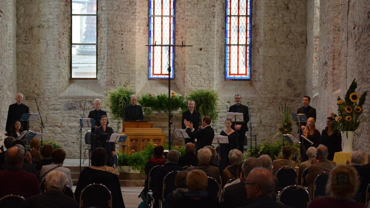 In einem Kirchenraum, der mit vielen Sonnenblumen geschmückt ist, stehen Musiker und Sängerinnen und bedanken sich beim Publikum, das verteilt im Raum sitzt und Beifall spendet. In einem Kirchenraum, der mit vielen Sonnenblumen geschmückt ist, stehen Musiker und Sängerinnen und bedanken sich beim Publikum, das verteilt im Raum sitzt und Beifall spendet.
