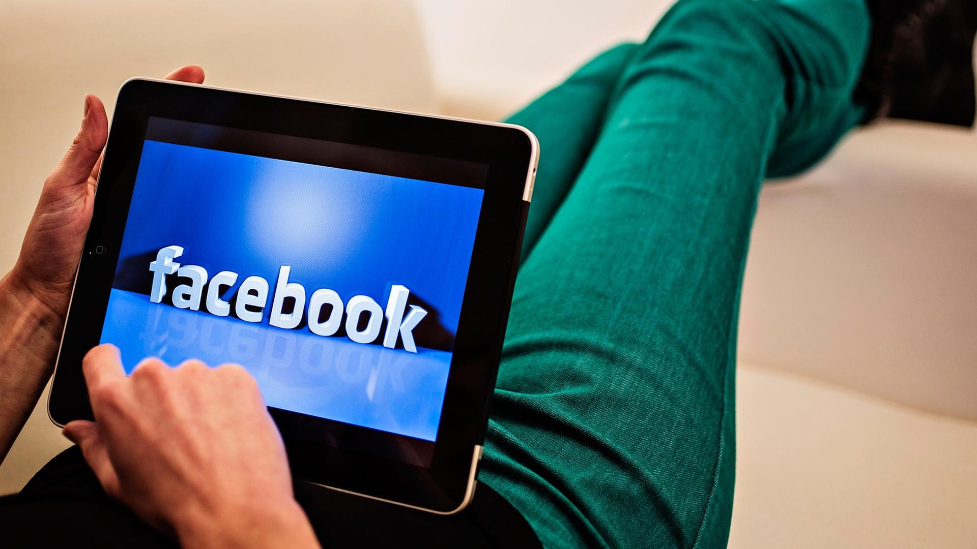 Ein Internetnutzer hat ein Tablet auf dem Schoß, darauf ist eine Facebook-Illustration zu sehen. Seine Beine sind über eine Sofalehne geschwungen. Ein Internetnutzer hat ein Tablet auf dem Schoß, darauf ist eine Facebook-Illustration zu sehen. Seine Beine sind über eine Sofalehne geschwungen.