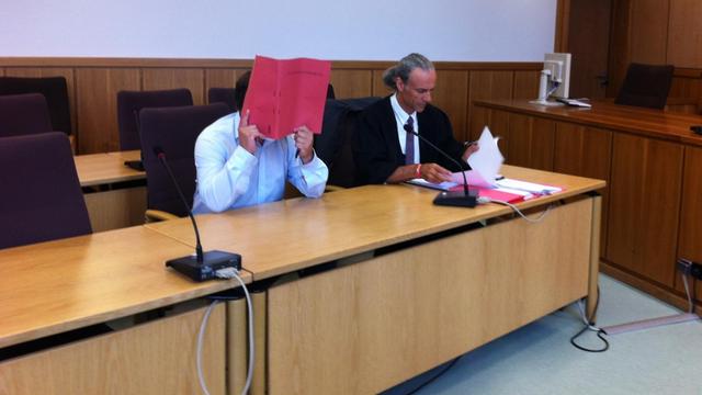 Der Angeklagte und sein Verteidiger im Gerichtssaal. Der Angeklagte bedeckt sein Gesicht mit einer Mappe.