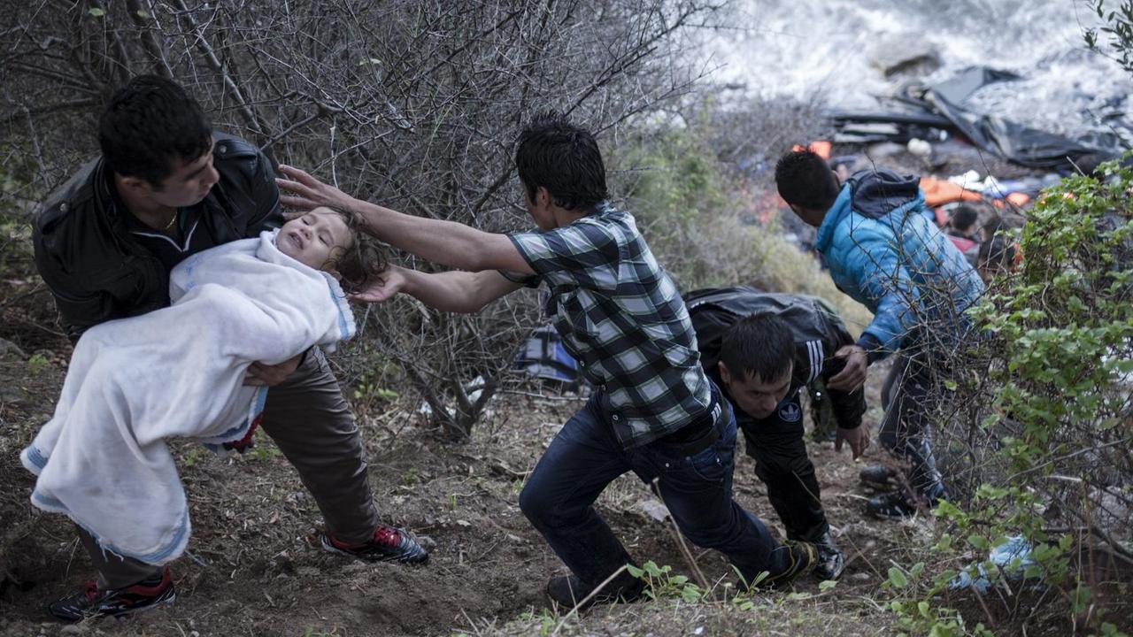 Ein Mann übergibt ein Kind an einen anderen, während sie an einer Stelle der nördlichen Küste der griechischen Insel Lesbos hochklettern. Am Wasser sieht man ein Schlauchboot, mit dem die Flüchtlinge angekommen sind. Ein Mann übergibt ein Kind an einen anderen, während sie an einer Stelle der nördlichen Küste der griechischen Insel Lesbos hochklettern. Am Wasser sieht man ein Schlauchboot, mit dem die Flüchtlinge angekommen sind.