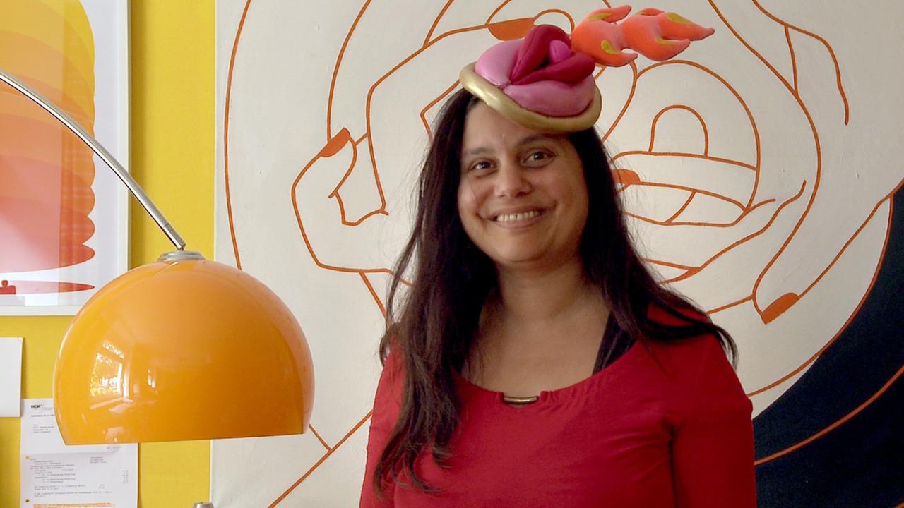 Mithu Sanyal trägt einen vulvös geformten Kopfschmuck und lacht in die Kamera. Mithu Sanyal trägt einen vulvös geformten Kopfschmuck und lacht in die Kamera.