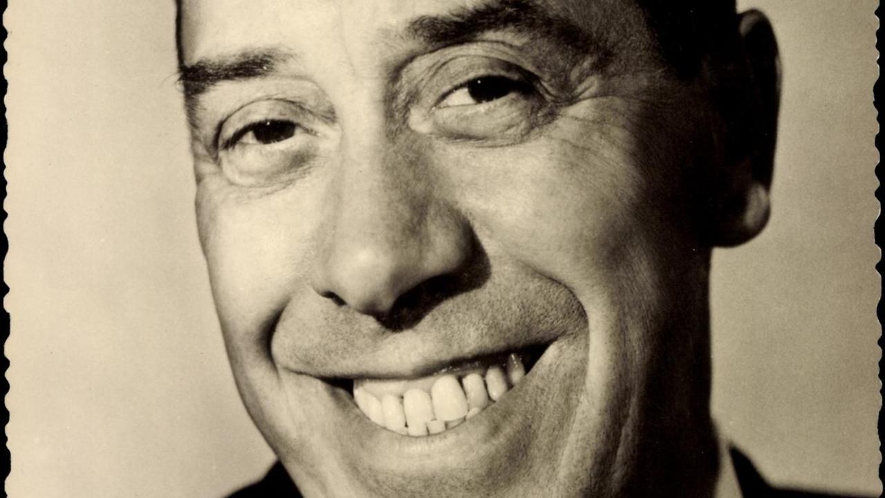 Ein schwarz-weißes Foto zeigt den Schauspieler und Komiker Fernandel in schwarzem Jackett, weißem Hemd und Krawatte breit in die Kamera grinsend Ein schwarz-weißes Foto zeigt den Schauspieler und Komiker Fernandel in schwarzem Jackett, weißem Hemd und Krawatte breit in die Kamera grinsend