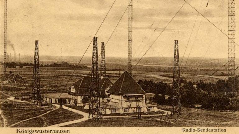 Eine historische Postkarte zeigt einige Gebäude, zwischen denen hohe Masten stehen, die über lang gespannte Seile gesichert sind. Eine historische Postkarte zeigt einige Gebäude, zwischen denen hohe Masten stehen, die über lang gespannte Seile gesichert sind.