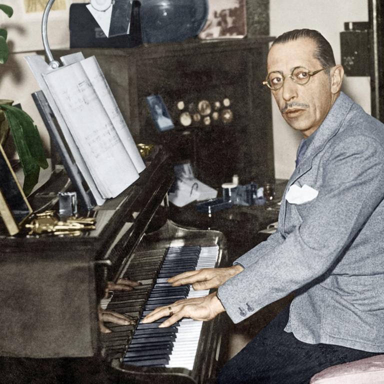 Ein Mann mit streng zurückgekämmten Haaren sitzt am Klavier und blickt durch eine runde Brille nach links in die Kamera. Er trägt hellgraues Jackett mit weißem Einstecktuch und schwarze Cordhose.