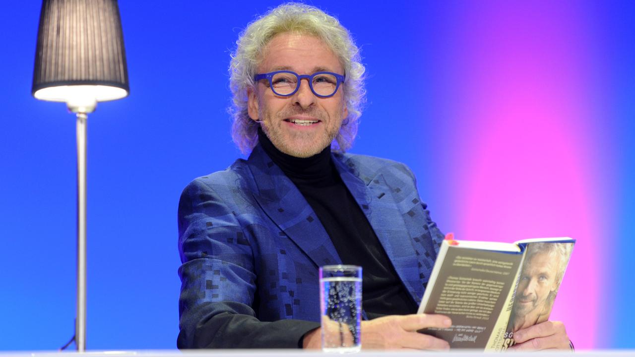 Thomas Gottschalk sitzt mit Brille in einem Fernsehstudio vor blau-violetten Hintergrund unter einer Stehlampe und hält ein Buch in der Hand. Vor ihm steht ein Wasserglas. Thomas Gottschalk sitzt mit Brille in einem Fernsehstudio vor blau-violetten Hintergrund unter einer Stehlampe und hält ein Buch in der Hand. Vor ihm steht ein Wasserglas.