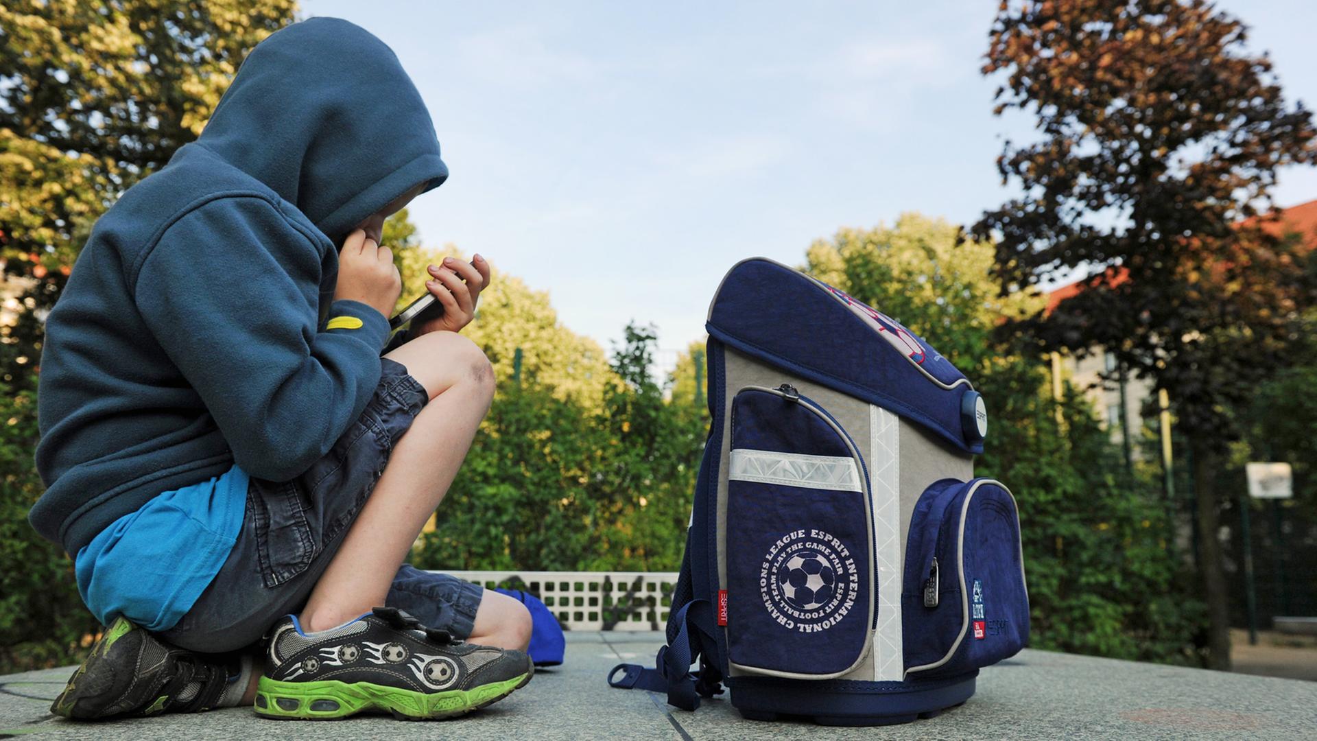 Ein Junge sitzt mit seinem Schulranzen auf einer Tischtennisplatte und spielt auf einem Smartphone. Ein Junge sitzt mit seinem Schulranzen auf einer Tischtennisplatte und spielt auf einem Smartphone.