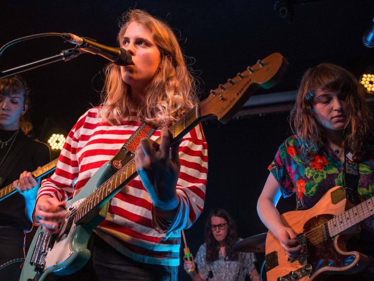 Die Musikerin Marika Hackman steht mit ihrer Gitarre auf der Bühne und singt. Sie wird von zwei weiteren Musikerinnen unterstützt.