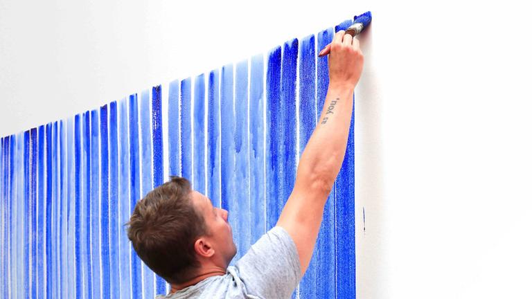 Jeppe Hein malt mit blauer Farbe und einem breiten Pinsel vertikale Linien auf eine Wand. Jeppe Hein malt mit blauer Farbe und einem breiten Pinsel vertikale Linien auf eine Wand.